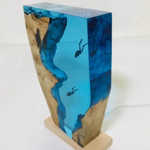 K&ouml;nnte beinhalten: Eine einzigartige Lampe mit rechteckigem Design, die einen klaren, blauen Harzmittelteil mit Tauchern und Seiten aus Naturholz aufweist. Die Lampe steht auf einem Holzsockel und hat ein Netzkabel.