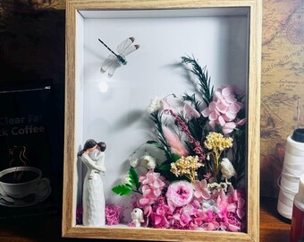 May include: A framed shadow box with a floral arrangement. The arrangement includes pink and white flowers, greenery, and a dragonfly figurine. A small figurine of a couple embracing and a small white bunny are also included.