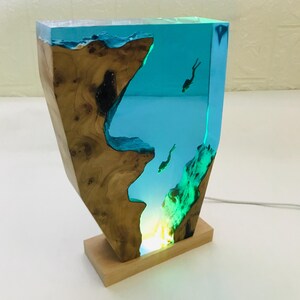 K&ouml;nnte beinhalten: Dekorative Tischlampe mit Holz- und Harzdesign. Die Lampe hat einen Holzsockel und Seiten mit einem transparenten Harz in der Mitte, das eine Unterwasserszene mit Tauchern darstellt. Das Harz ist leuchtend blau, und die Lampe strahlt ein weiches, warmes Licht aus.