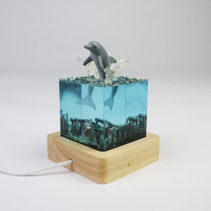May include: A gray dolphin figurine leaping out of a blue resin cube with a wooden base. The cube contains a miniature underwater scene with a light inside.
