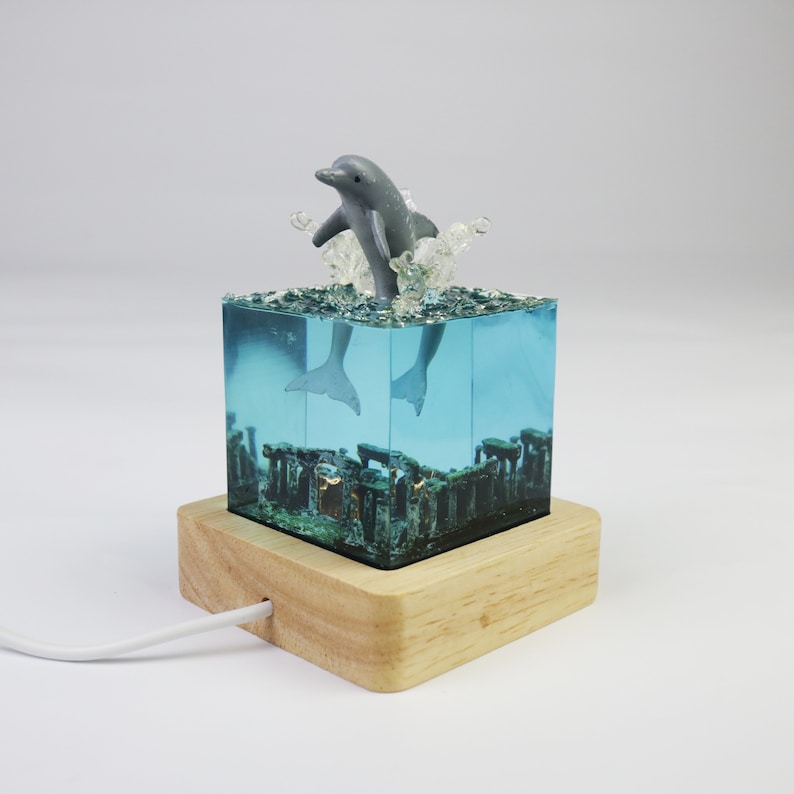 May include: A gray dolphin figurine leaping out of a blue resin cube with a wooden base. The cube contains a miniature underwater scene with a light inside.