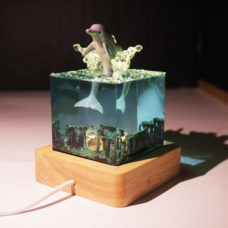 May include: A clear cube with a dolphin figurine inside, surrounded by green and brown resin. The cube is on a wooden base with a white cord attached. The dolphin is jumping out of the water, with its tail and fins visible.