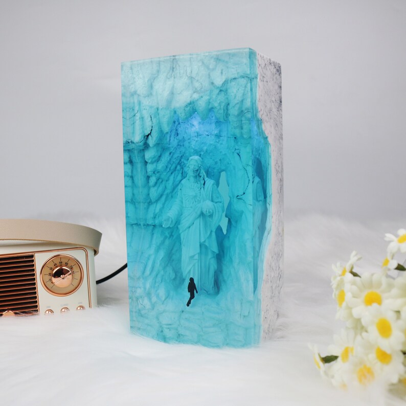 May include: A blue resin sculpture of a person standing in a cave with a white border. The sculpture is lit from behind, creating a glow effect.