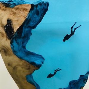 K&ouml;nnte beinhalten: Ein einzigartiges Kunstwerk aus Kunstharz mit einem Holzsockel und blauem Kunstharz, das Wasser simuliert. Zwei Taucher-Silhouetten sind in dem blauen Kunstharz schwebend, was Tiefe erzeugt. Das Holz hat eine nat&uuml;rliche, hellbraune Farbe.