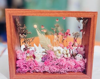 May include: A decorative wooden framed shadow box featuring a miniature garden scene. The scene includes pink moss, dried flowers, and small white bunny figurines. The frame is a warm brown color.