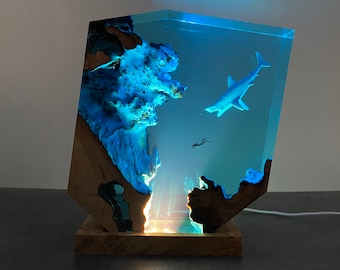 May include: A blue resin sculpture of a shark and a diver, set in a wooden base with a light inside. The resin is clear and the shark and diver are visible through the resin.