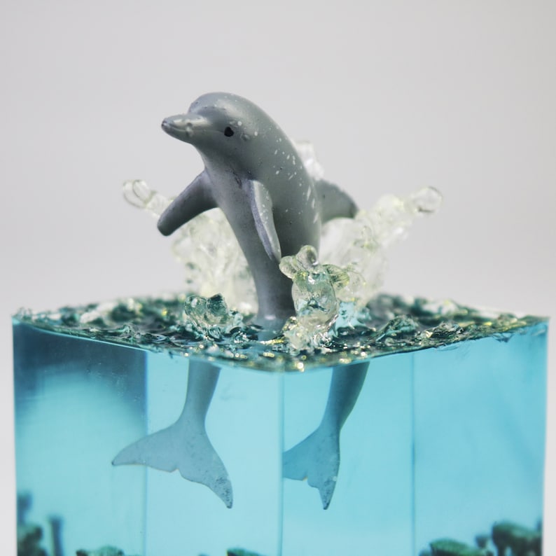 May include: A gray dolphin figurine leaping out of a clear blue resin block, creating a wave effect. The dolphin is positioned mid-jump, with its tail and fins visible below the waterline.