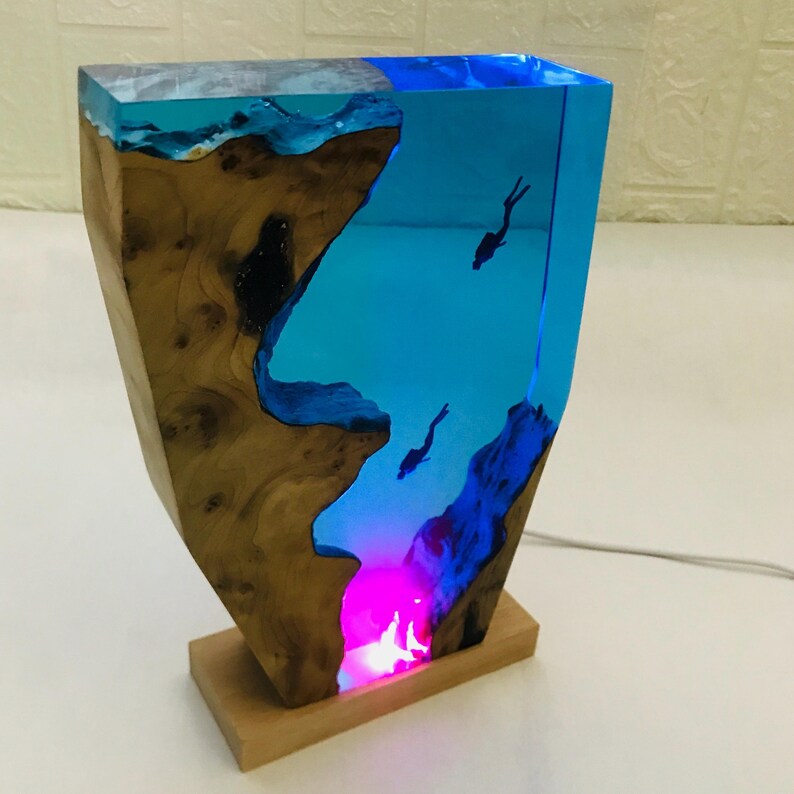 K&ouml;nnte beinhalten: Dekorative Lampe mit Holz- und Harzdesign. Die Lampe hat einen Holzsockel und einen transparenten Harzabschnitt mit blauem Farbton, der Taucher zeigt. Die Lampe strahlt ein farbenfrohes Leuchten aus.
