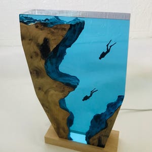K&ouml;nnte beinhalten: Dekorative Lampe mit Holz- und Harzdesign. Die Lampe hat eine rechteckige Form mit einem Holzsockel. Das Harz ist klarblau, mit zwei Tauchersilhouetten. Das Holz hat eine nat&uuml;rliche, braune Maserung. Ein einzigartiger Einrichtungsgegenstand.