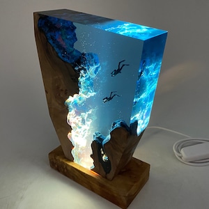 May include: A blue resin lamp with a wooden base, featuring a scene of divers swimming in a blue ocean. The lamp is lit, casting a warm glow.
