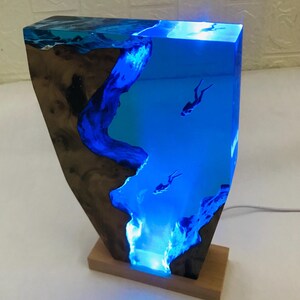 K&ouml;nnte beinhalten: Harzlampe mit einer Unterwasserszene und Tauchern. Die Lampe hat einen Holzsockel und eine rechteckige Form. Das blaue Harz ist beleuchtet und erzeugt einen Leuchteffekt. Die Lampe ist etwa 30 cm hoch.