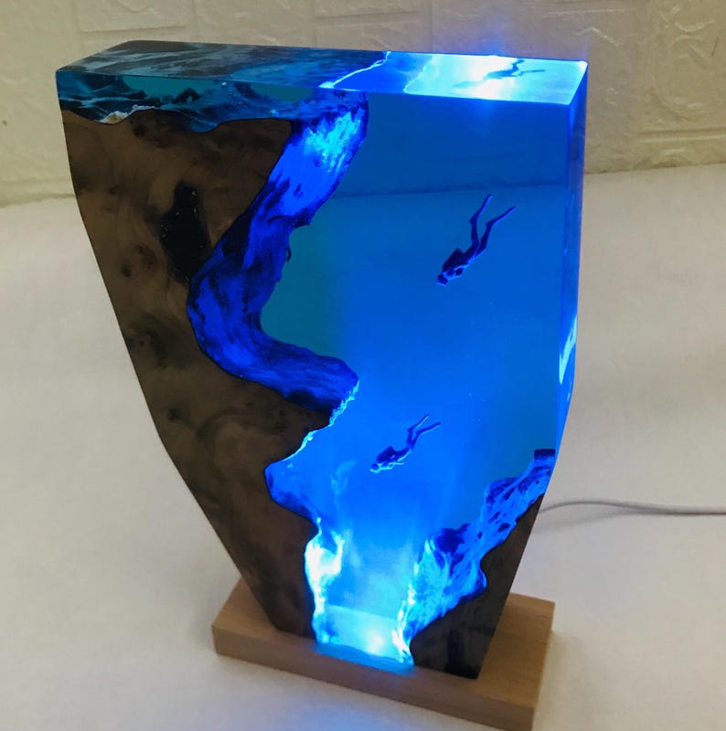 K&ouml;nnte beinhalten: Harzlampe mit einer Unterwasserszene und Tauchern. Die Lampe hat einen Holzsockel und eine rechteckige Form. Das blaue Harz ist beleuchtet und erzeugt einen Leuchteffekt. Die Lampe ist etwa 30 cm hoch.
