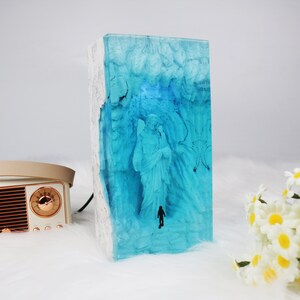 May include: A blue resin sculpture of a person standing in a cave. The sculpture is encased in clear resin and has a white base.