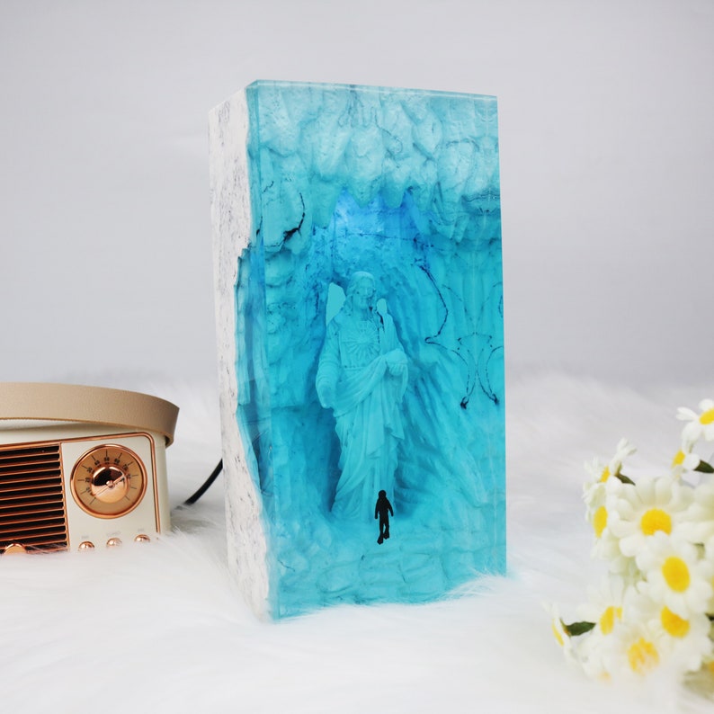 May include: A blue resin sculpture of a person standing in a cave. The sculpture is encased in clear resin and has a white base.
