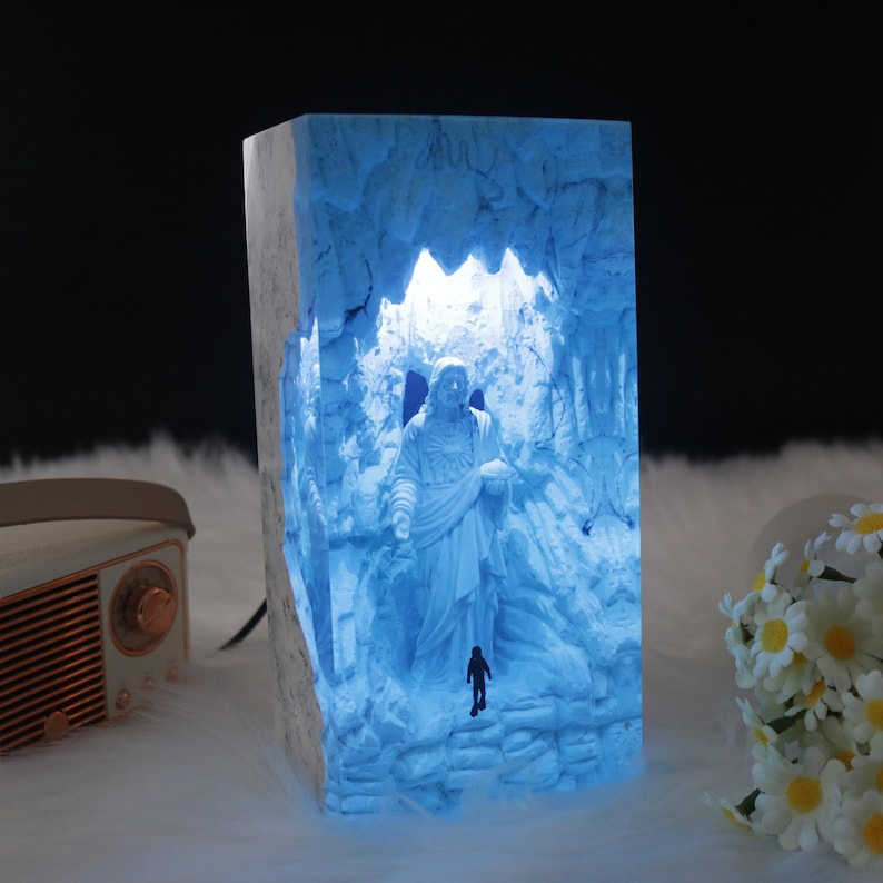 May include: A blue resin lamp with a 3D sculpture of a person standing in a cave. The lamp is lit from inside, casting a blue glow on the sculpture.