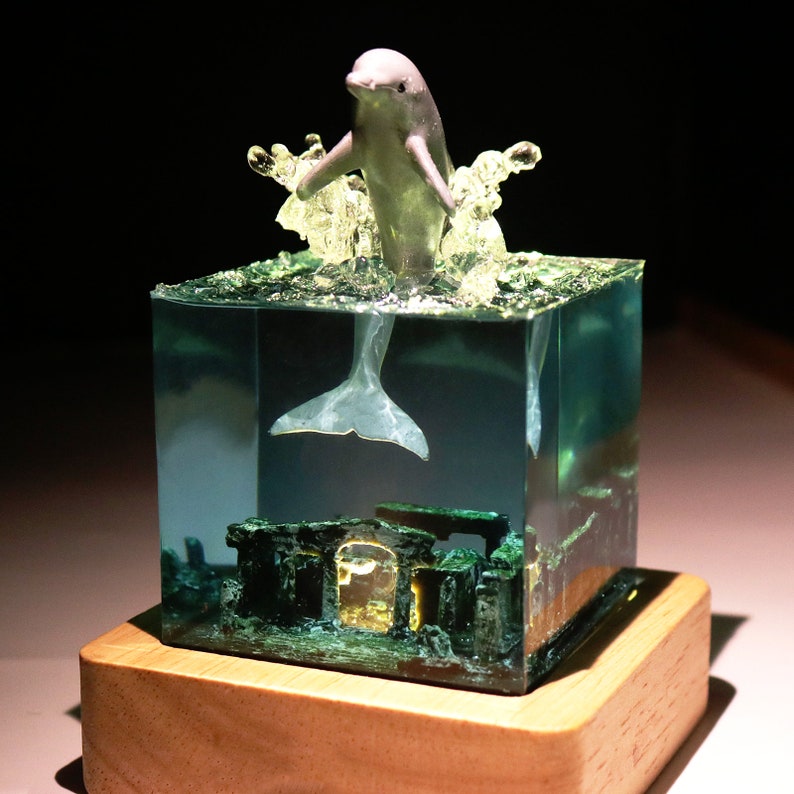 May include: A clear resin cube with a small white dolphin figurine jumping out of the water. The dolphin's tail is visible below the waterline. The cube is sitting on a wooden base.