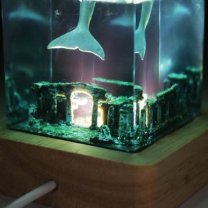 May include: A clear resin lamp with a white dolphin figurine swimming inside. The base of the lamp is made of wood and has a light cord.