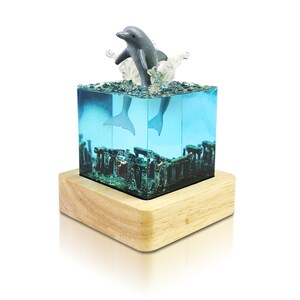May include: A gray dolphin figurine leaping out of a clear blue resin cube with a wooden base. The cube contains a miniature underwater scene with coral and seaweed.