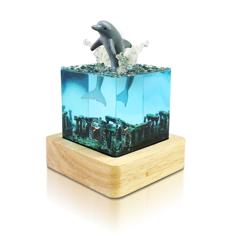 May include: A gray dolphin figurine leaping out of a clear blue resin cube with a wooden base. The cube contains a miniature underwater scene with coral and seaweed.