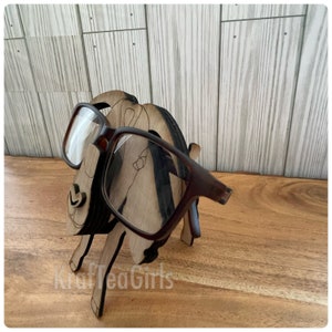 May include: A wooden eyeglasses holder shaped like a goat with brown glasses resting on the holder. The goat is standing on a wooden surface.