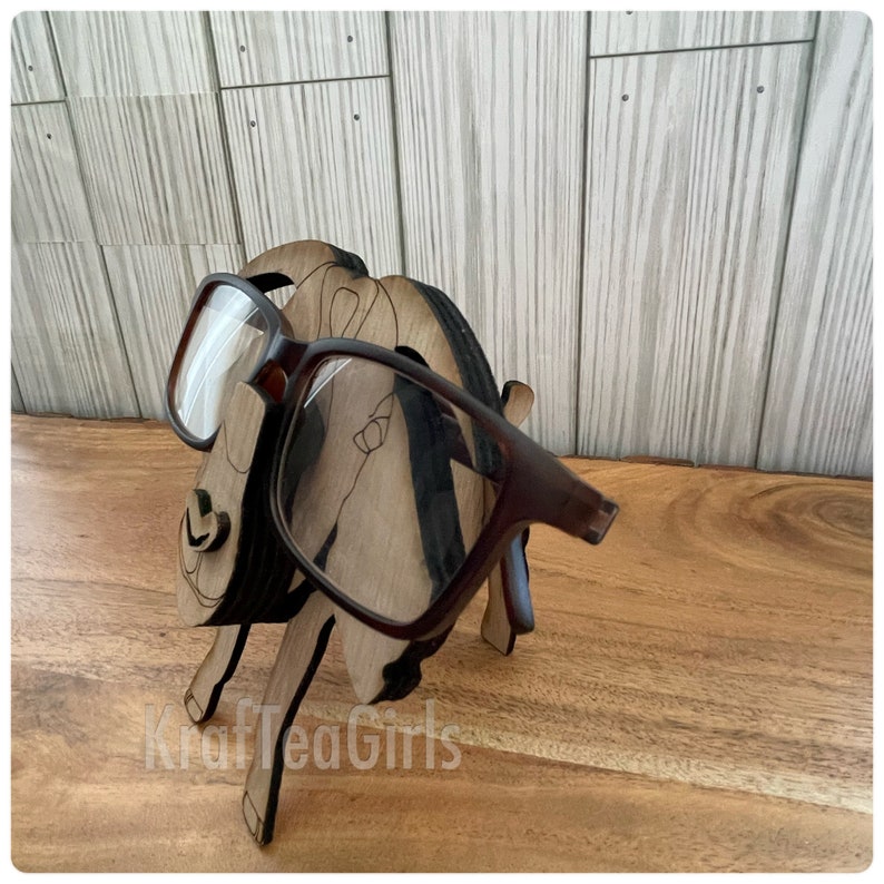 May include: A wooden eyeglasses holder shaped like a goat with brown glasses resting on the holder. The goat is standing on a wooden surface.