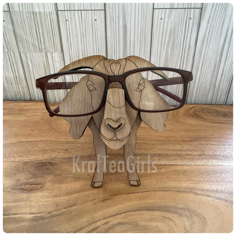 May include: A wooden goat figurine wearing brown glasses. The goat is standing on a wooden surface. The text "KrafTeaGirls" is visible on the surface.