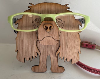 May include: A wooden dog figurine with a light brown finish, wearing green glasses. The dog has a long, flowing mane and a friendly expression.
