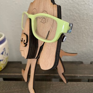 May include: Wooden dog figurine with a light brown finish, standing on a wooden surface. The dog is wearing green glasses.