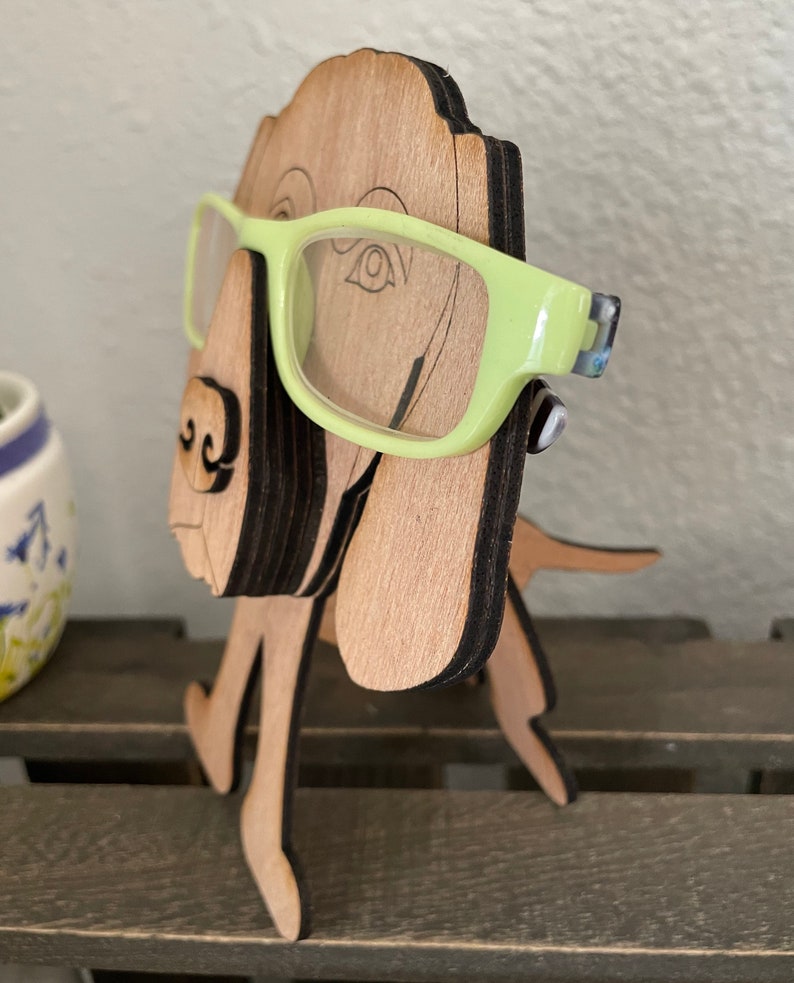 May include: Wooden dog figurine with a light brown finish, standing on a wooden surface. The dog is wearing green glasses.