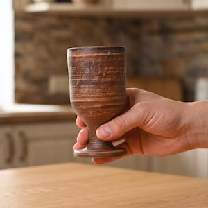 May include: A brown clay goblet with a smooth, rounded bowl and a sturdy, flared base. The surface features horizontal striations and a natural, earthy texture with variations in color ranging from dark brown to lighter reddish-brown tones.