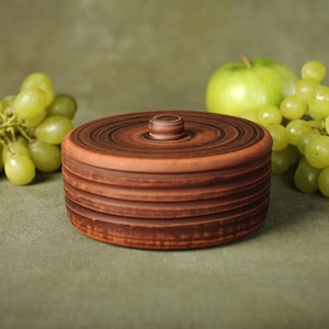 May include: A brown ceramic container with a lid, sitting on a green surface with green grapes and a green apple.