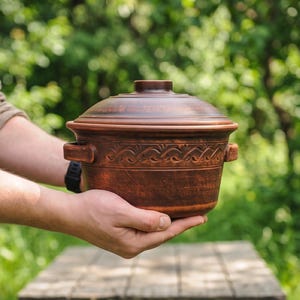 May include: A round, brown clay cooking pot with a matching lid and two side handles. The exterior of the pot features a decorative embossed wave pattern around the center. The surface has a smooth, earthy texture with natural color variations.