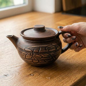 Könnte beinhalten: Braune Keramik-Teekanne mit Deckel und Henkel, mit einem floralen Design, das in die Oberfläche geschnitzt ist. Die Teekanne steht auf einer Holzoberfläche.