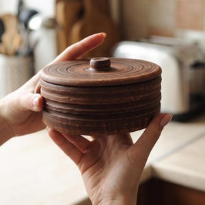 May include: A round, dark brown clay container with a matching lid. The exterior features horizontal ridges and a smooth, circular knob on top. The surface has a natural, earthy texture with subtle variations in colour throughout the clay material.