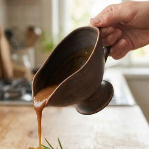 May include: A brown gravy boat pours gravy over a plate of roasted meat, potatoes, and carrots. The gravy boat is made of a dark brown material and has a handle. A sprig of rosemary is on the plate. The food is on a light brown plate.