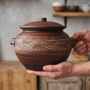 May include: A round, dark brown clay pot with a matching lid and two small side handles. The exterior features a horizontal band of carved, wave-like leaf patterns encircling the middle section, with a smooth finish on the base and lid.