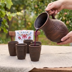 May include: A dark brown clay pitcher with an embossed wheat stalk design pouring dark purple liquid into one of four matching clay tumblers. Each tumbler features a decorative band around the rim, set on a light-colored linen tablecloth.