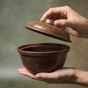 Peut inclure: Un bol en céramique marron avec un couvercle assorti soulevé. Le bol est rond avec un bord légèrement évasé. Le couvercle a une petite poignée. Le bol est tenu dans les mains d'une personne. Diamètre du bol : environ 15 cm.