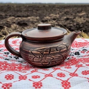 May include: A brown clay teapot with a floral design sits on a white tablecloth with red and black embroidery. The teapot is on a bed of dry grass.