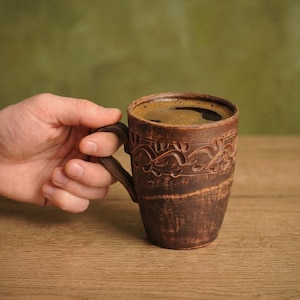 May include: A brown ceramic mug filled with dark coffee. The mug has a handle and is decorated with carved floral patterns. The mug is held by a hand, and the background is a blurred green and brown.