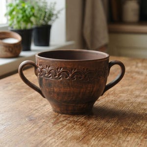 May include: A dark brown clay bowl with two curved handles on opposite sides. The exterior features a decorative embossed pattern of repeating leaf-like shapes encircling the upper rim. The surface has a natural, matte, and textured finish.
