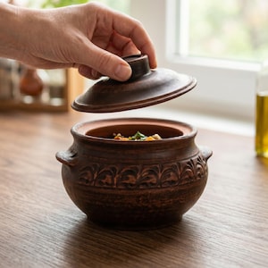 May include: A brown ceramic cooking pot with a lid being lifted, revealing a glimpse of the food inside. The pot has decorative carvings and small handles. The pot is sitting on a wooden surface.
