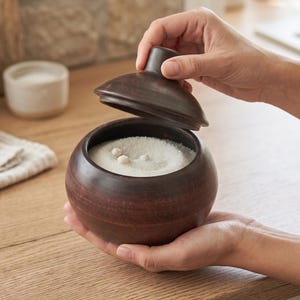 May include: A round, dark brown clay bowl with a matching lid held slightly open. The bowl contains a white granular substance with a few small, round, light-colored beads resting on the surface. The container has a smooth, matte finish.