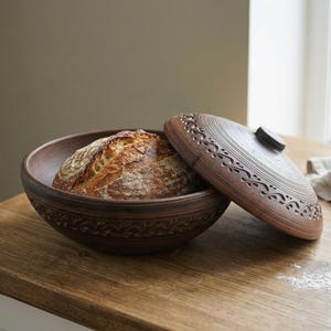 May include: A brown ceramic bread bowl with a decorative lid, holding a loaf of bread. The lid is partially open, revealing the bread. The bowl and lid have carved patterns. The bread has a golden crust.