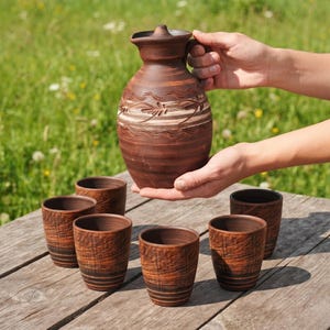 May include: A brown clay pitcher with a lid and a decorative carved leaf pattern around the centre, accompanied by five matching brown clay cups. The pitcher and cups feature a textured, rustic finish with subtle variations in the dark brown clay.