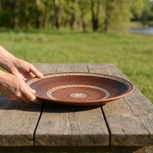 Pode incluir: Uma taça de terracota redonda e rasa com um acabamento liso castanho escuro. A taça apresenta duas faixas circulares concêntricas com padrões geométricos intrincados em cor creme, uma ao longo da borda interna e outra no centro da base.