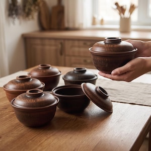 May include: A set of dark brown clay bowls with matching lids. The bowls have a rounded shape and a smooth, earthy texture. One bowl is held in the air, while several others are arranged on a wooden surface, some with lids on and one open.