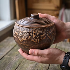 Peut inclure: Un pot en argile brun foncé de forme ronde avec un couvercle assorti. L'extérieur présente un motif sculpté et texturé sur la section centrale, tandis que le couvercle comporte des crêtes circulaires concentriques. Le récipient a une base arrondie.