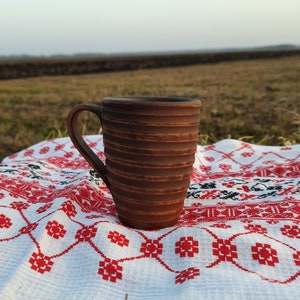 Könnte beinhalten: Eine braune Keramiktasse mit einem strukturierten, gerippten Design steht auf einer weißen Tischdecke mit einem roten und schwarzen Stickmuster. Die Tasse ist scharf, während der Hintergrund unscharf ist und ein Grasfeld und einen fernen Horizont zeigt.
