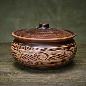Könnte beinhalten: Ein brauner Keramiktopf mit Deckel, der mit einem dekorativen geschnitzten Design aus wirbelnden Linien und Blättern verziert ist. Der Topf steht auf einer rustikalen Holzoberfläche.